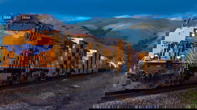 A train travels on train tracks. Behind the train are mountains.
