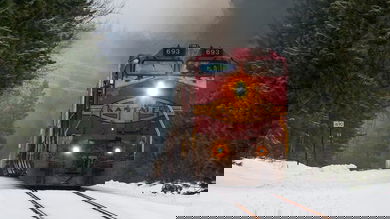 A photograph of a train with a headlight on. There is snow on the ground.
