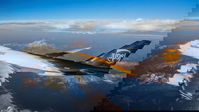 UJPS brown and white jumbo jet flies over mountains.
