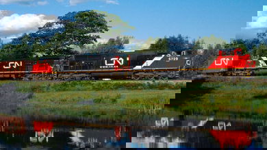 A photograph of a train traveling in a grassy field. A reflection of the train is on the water of a nearby pond.
