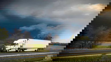 Tractor-trailer heading down a highway with dark storm cloud across the sky.