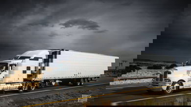 Tractor-trailer heading down highway with dark storm cloud across the sky.