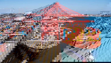 A big container ship stacked with boxes next to the wharf with big cranes and containers stacked on land.