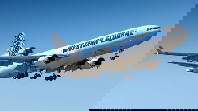 A white jet with three engines comes in for landing with wheels down against blue sky.