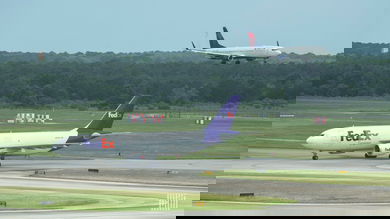 A white and purple FedEx plane on the tarmac as a Delta plane comes in for landing on another runway.