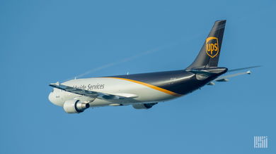 A UPS cargo plane, white in front and brown in rear, flying against blue sky, viewed from behind.