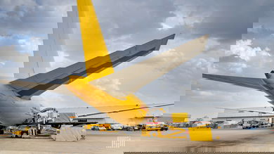 A mustard-yellow DHL plane loading cargo at an airport. View from behind the plane.