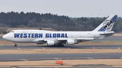 A white 747 jumbo jet with blue lettering: Western Global rolls down taxiway on a hazy day.