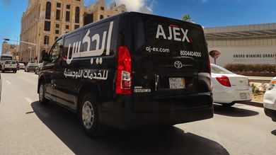 A black delivery van with Ajex Logistics logo driving in the city on a sunny day.