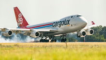 A red-tailed Cargolux jumbo jet touches down on a runway, with green grass in foreground.