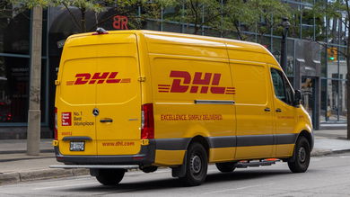 A mustard-colored DHL van with red lettering on a city street.