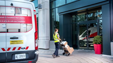 A mail carrier delivers packages from the van to an office building using a hand cart.