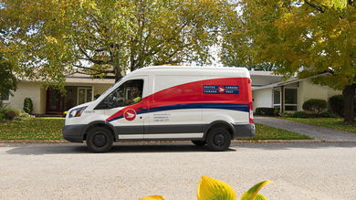 A Canada Post delivery van with red stripe drives through a suburban neighborhood.